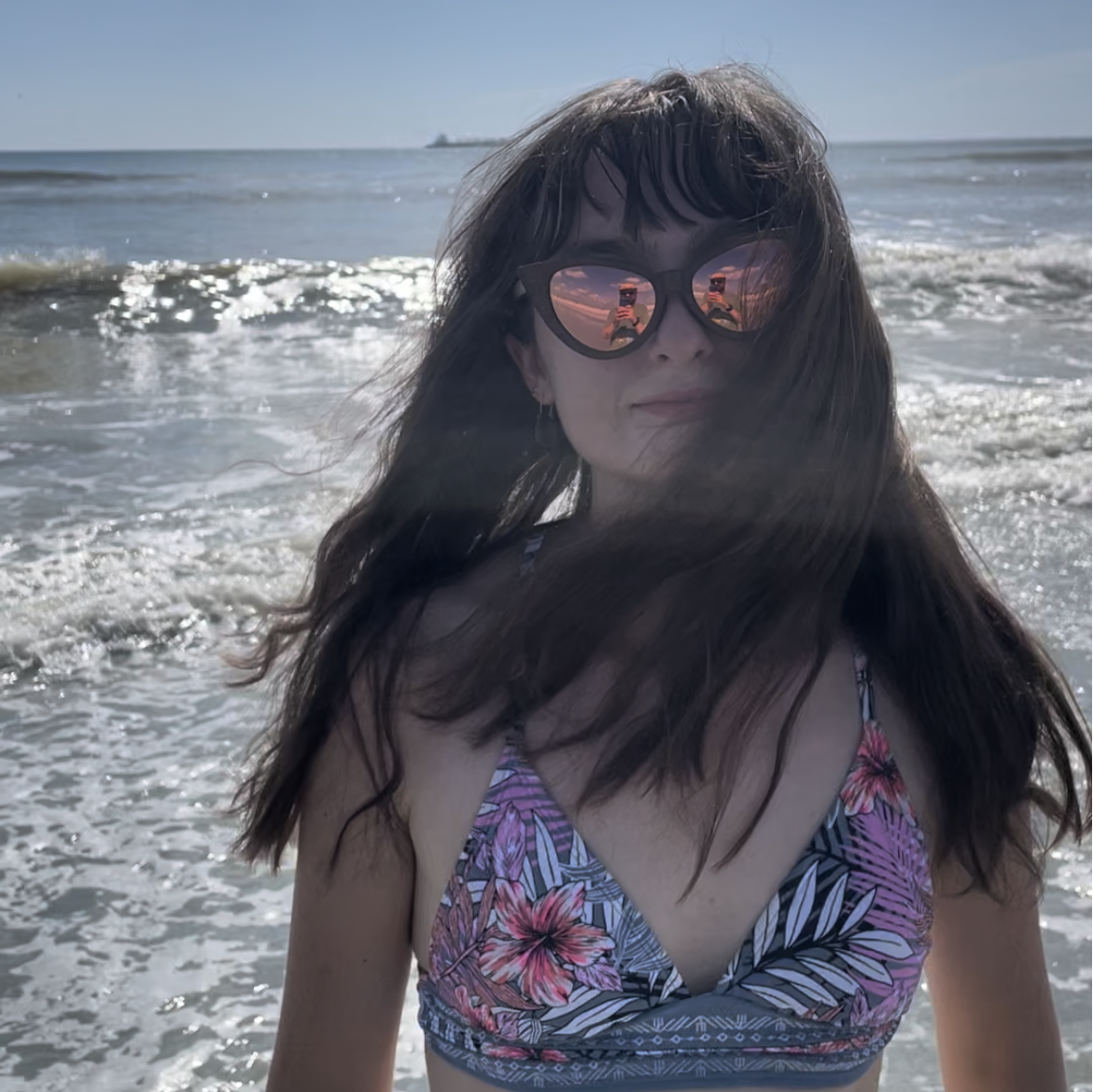 Woman wearing Ecochic Shades cat eye sunglasses in a colorful bikini and standing on a beach with ocean waves in the background.